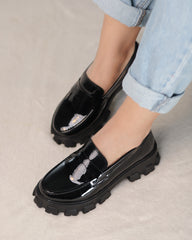 A pair of glossy black loafers with a sleek design, displayed on a person's feet.