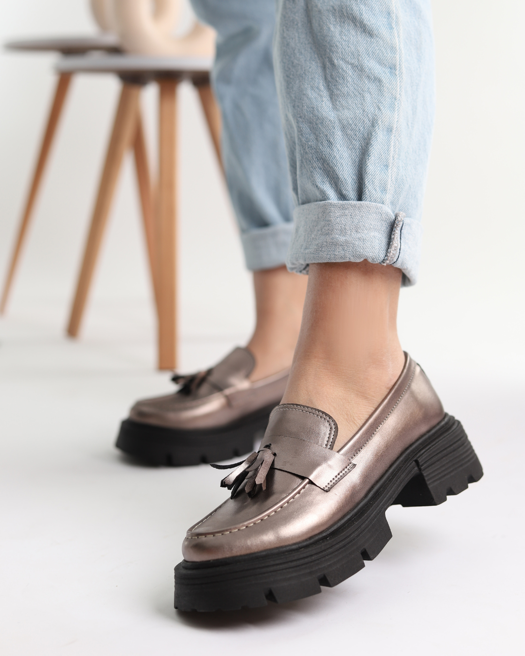 A person wearing metallic silver loafers with black tassels, paired with blue jeans, standing in a room with a wooden stool and a sculpture in the background.