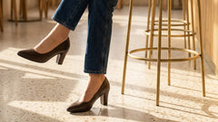Person sitting on a bar stool wearing blue jeans and black high heels in a bright room.