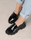 A pair of glossy black loafers with a sleek design, displayed on a person's feet.
