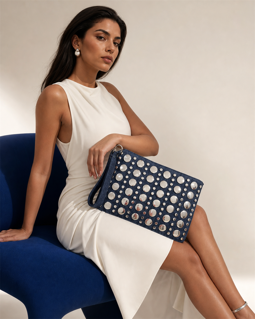 Denara studded denim clutch with seated female model on cobalt chair - creative view